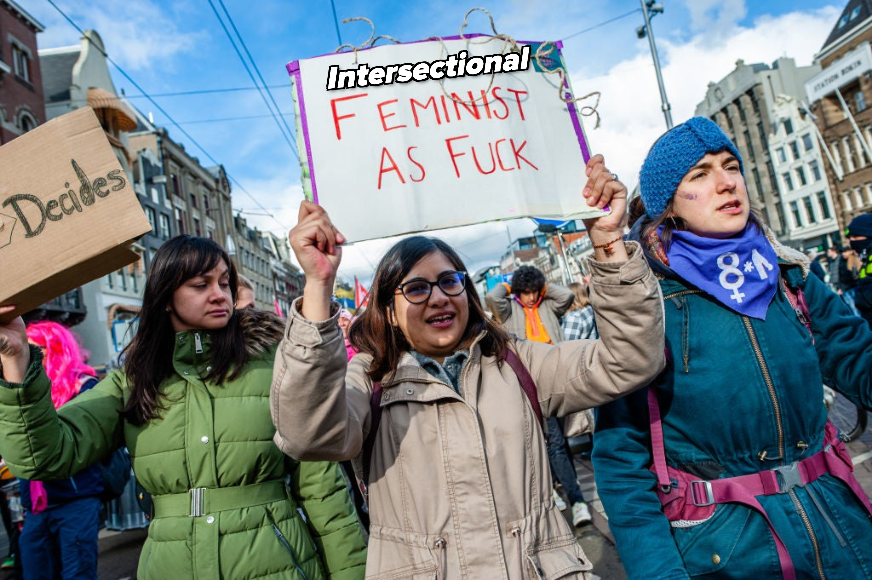 Three individuals at a rally holding a sign that reads &quot;Feminist as F***&quot; expressing advocacy for women&#x27;s rights