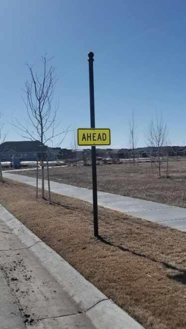 Traffic sign labeled &quot;AHEAD&quot; on a post beside a sidewalk with houses in the background