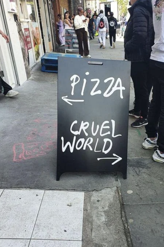 Signboard on a sidewalk with arrows pointing left to &quot;PIZZA&quot; and right to &quot;CRUEL WORLD.&quot;