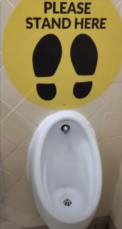 Sign above urinal reads &quot;PLEASE STAND HERE&quot; with footprints indicating where to stand
