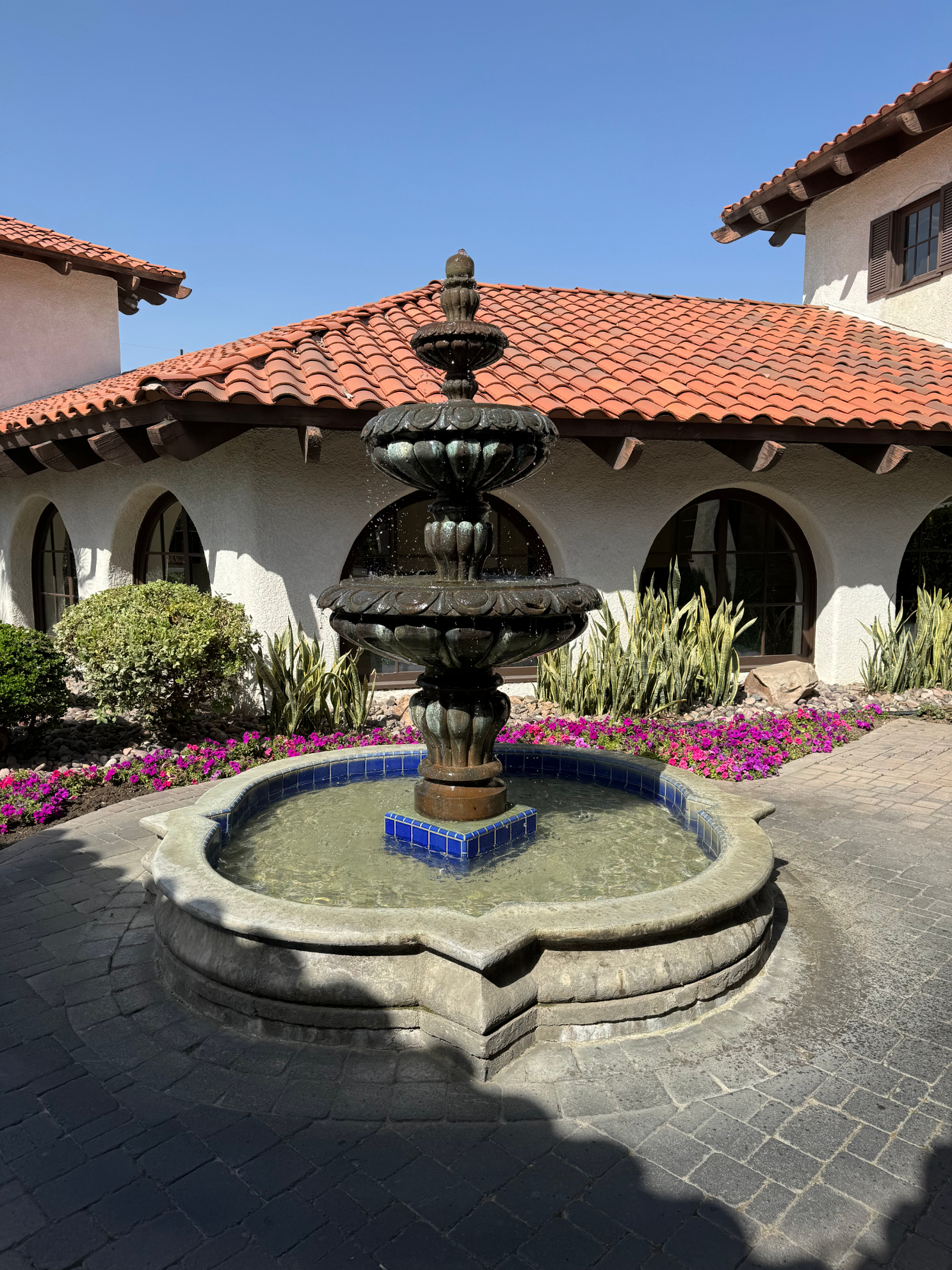 Multi-tiered fountain in front of a Spanish-style building, surrounded by purple flowers