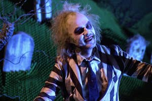 Character Beetlejuice in a striped suit gesturing, with a tombstone and eerie setting in the background