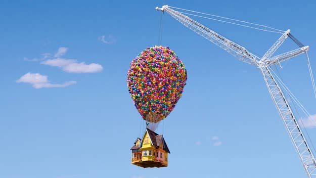 A house lifted by numerous balloons next to a crane against a blue sky
