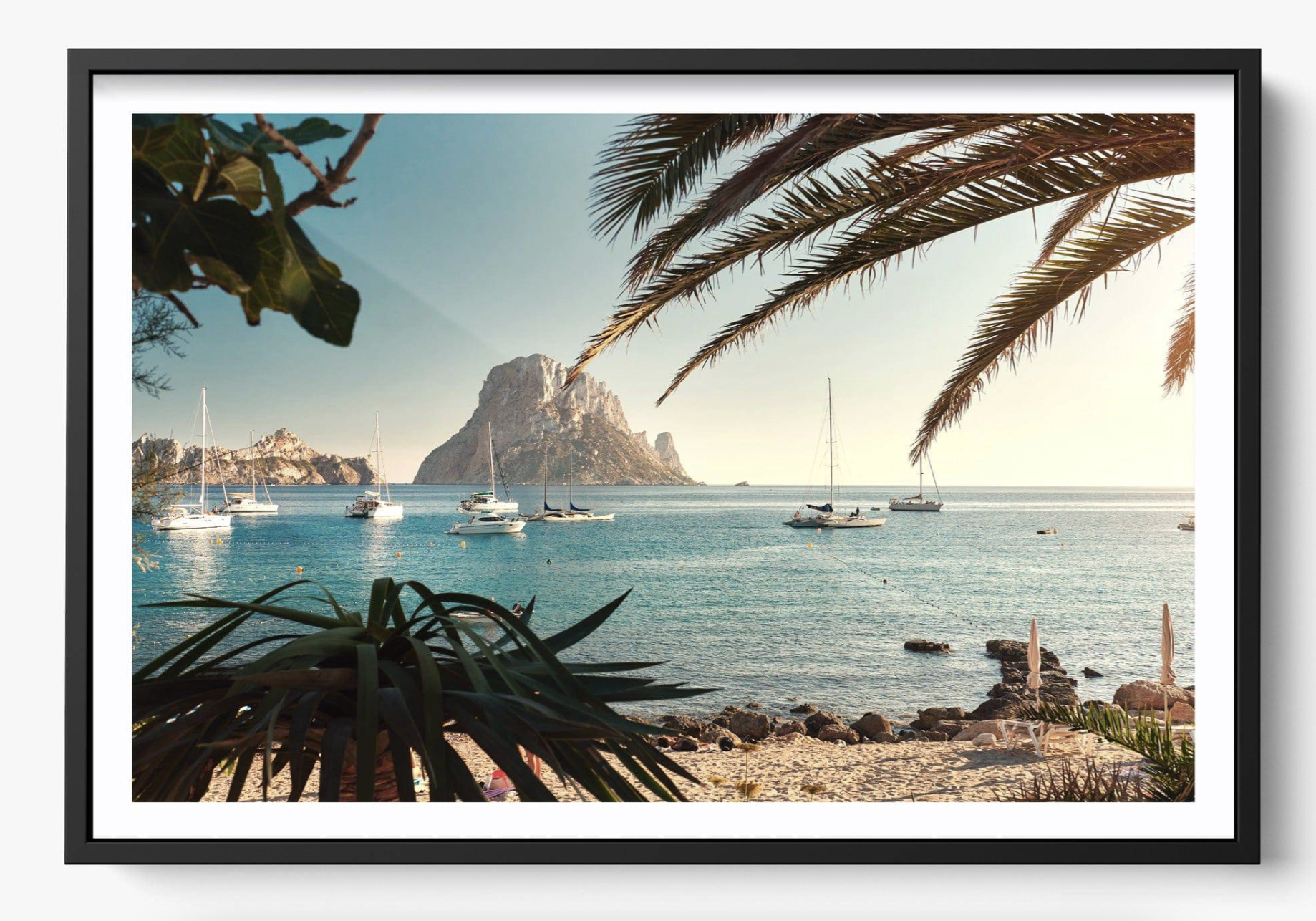 Scenic view of a calm bay with yachts and a prominent rocky outcrop in the distance, framed by palm leaves