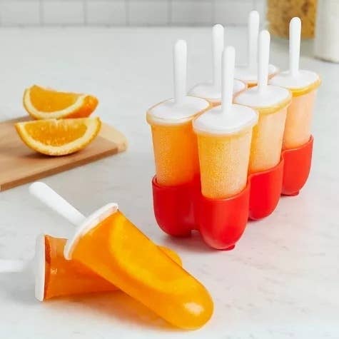 Homemade orange popsicles in a red mold with white sticks, some lying on a surface