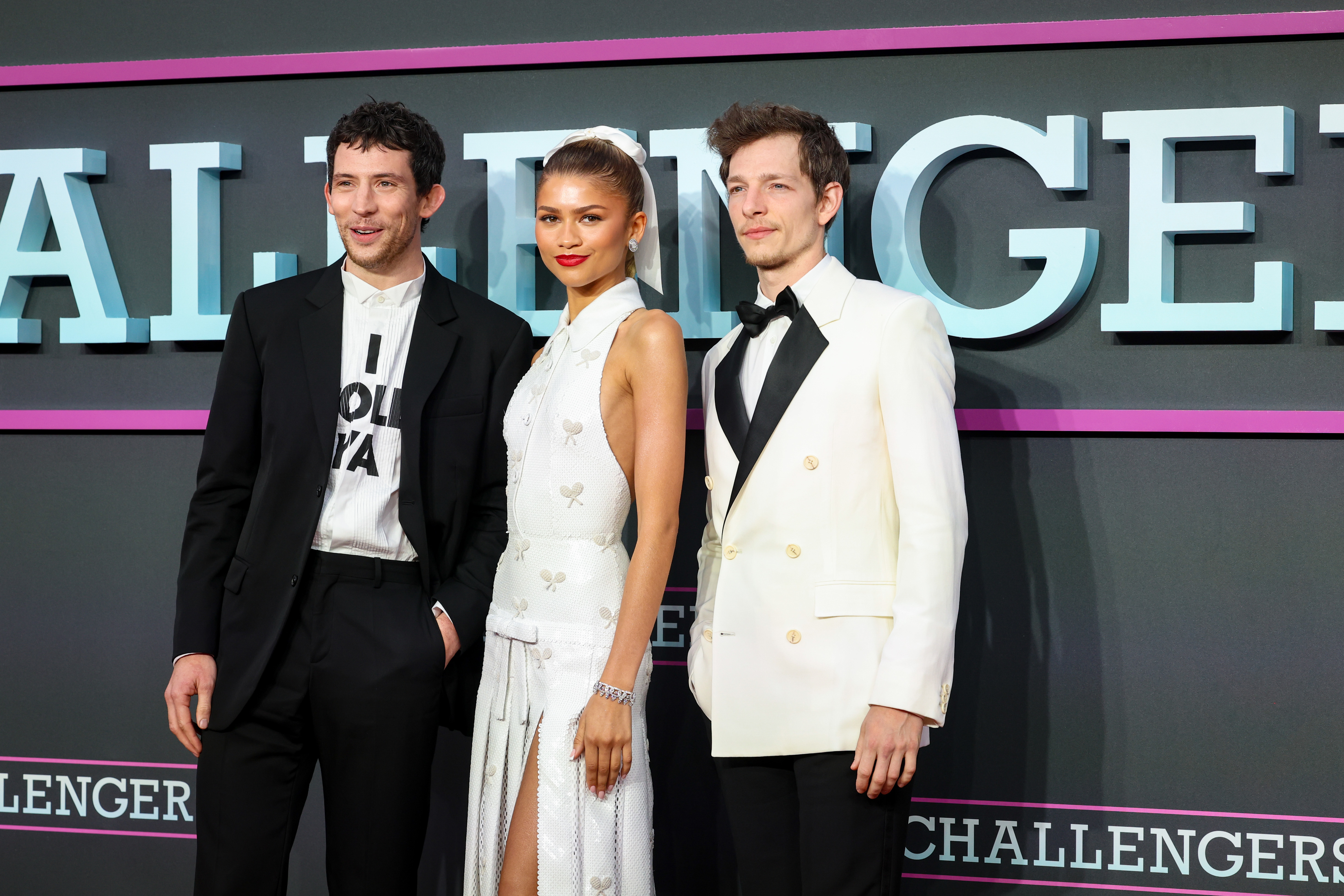 Three people posing for a photo; two men in suits with one woman between them, wearing a sleeveless dress, at a promotional event