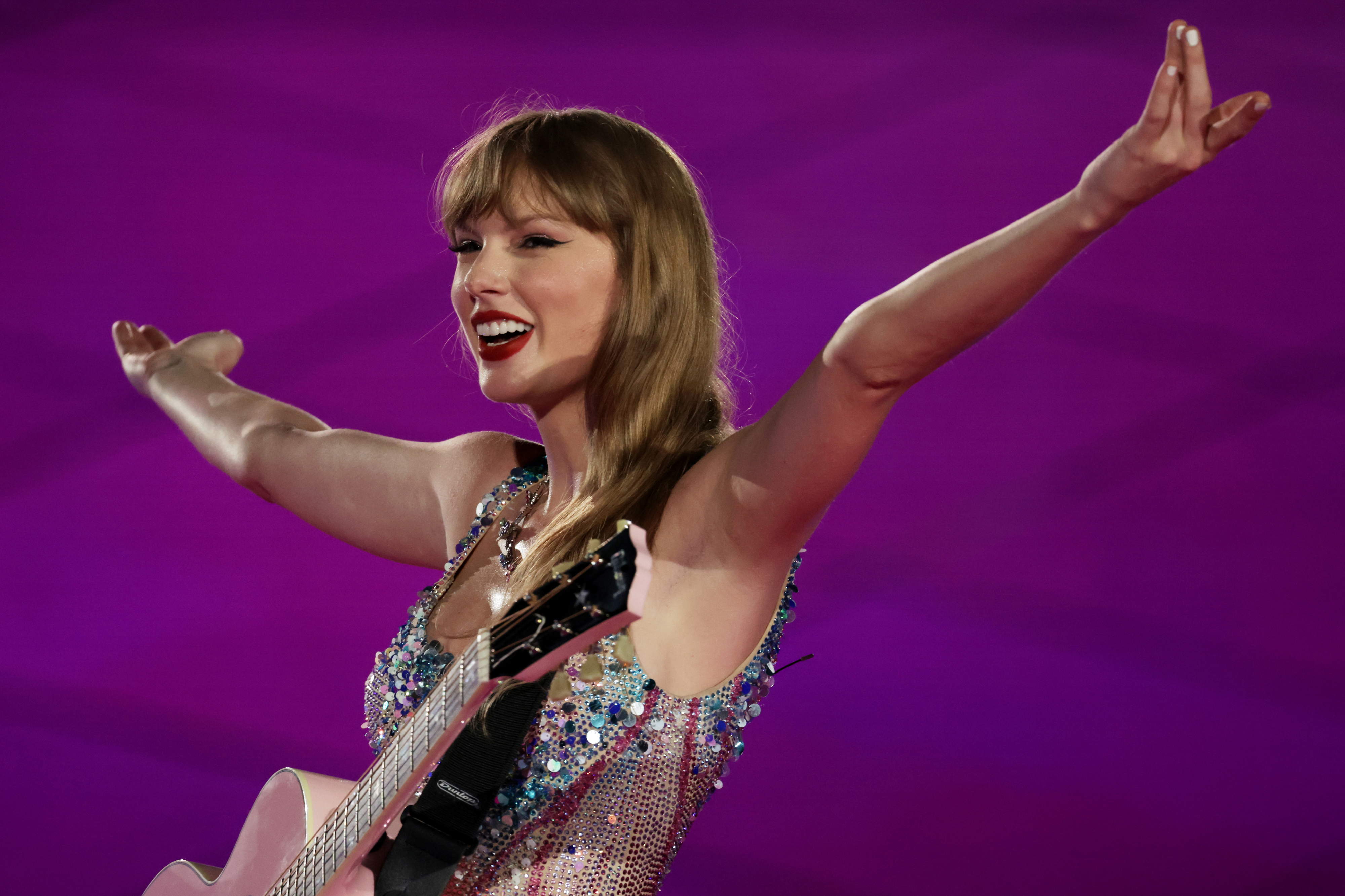 Taylor Swift performing with a guitar, wearing a sparkling dress, arms wide open