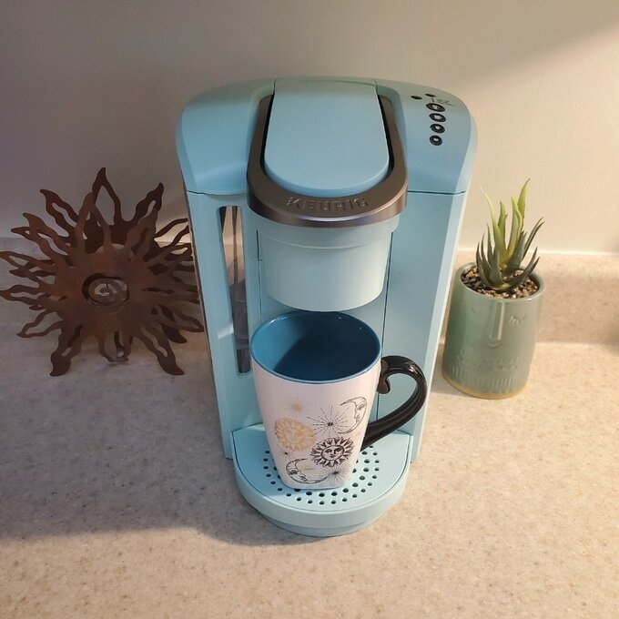 A Keurig coffee maker with a mug placed on the drip tray, next to a decorative item and a potted plant