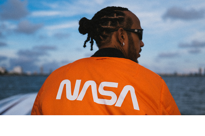 Person in orange NASA jacket facing away, looking at scenic sky with water in the background
