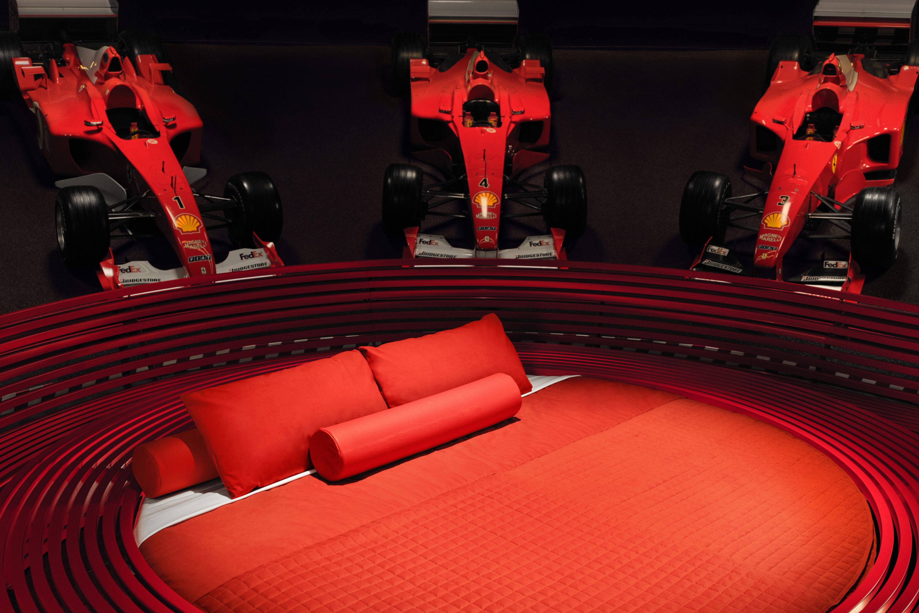 Two Ferrari Formula 1 cars flank a circular red couch in a room with concentric circles on the floor