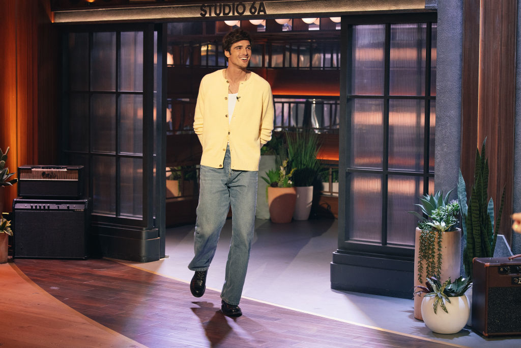 Person in a casual blazer and jeans enters a stage with "STUDIO 6A" sign overhead