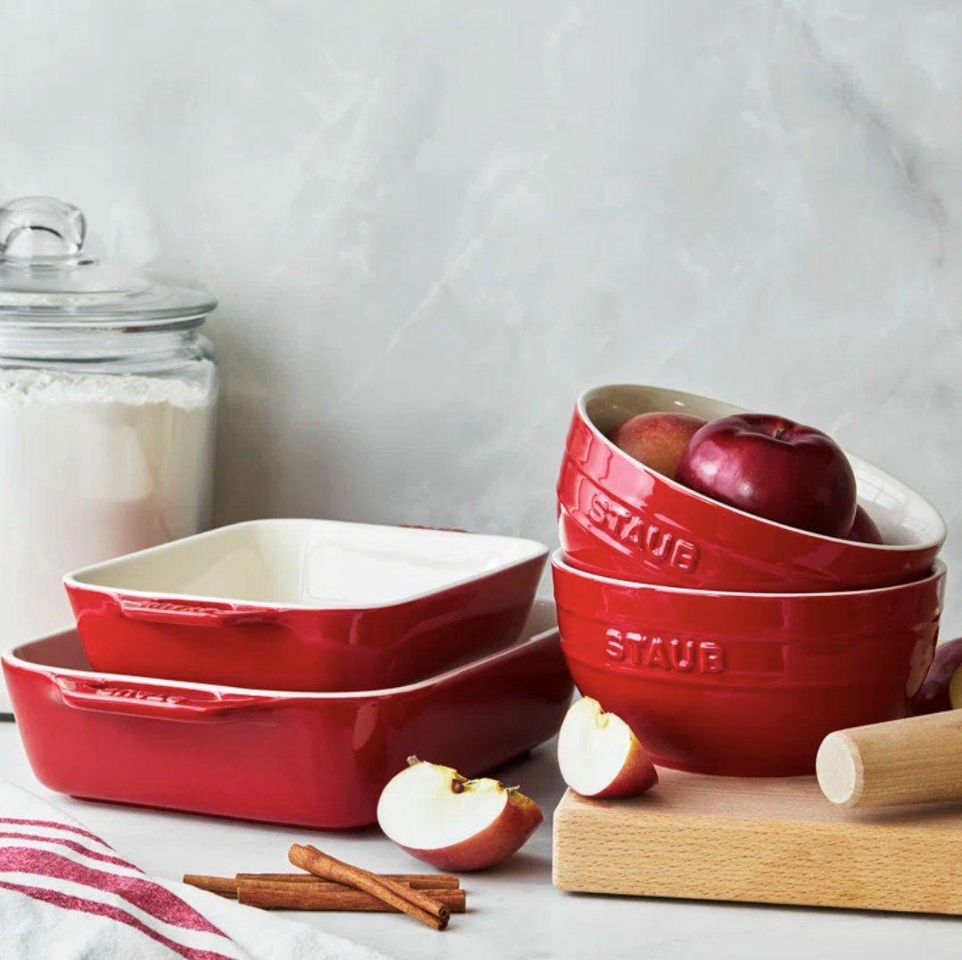 Four red Staub ceramic cookware pieces with apples and kitchen utensils on a counter