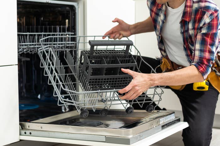 Person repairing an open dishwasher, hands on the racks, tool belt visible