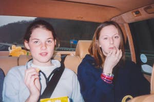Two individuals in a car; one is eating snacks and the other is applying makeup