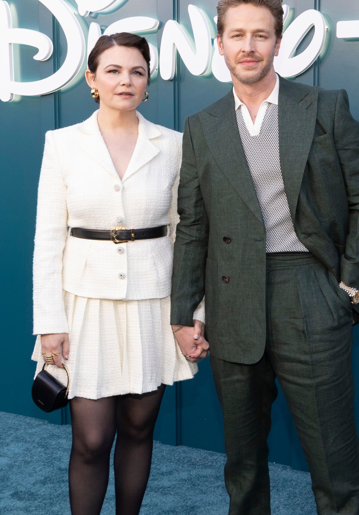 Two celebrities pose at a Disney+ event; one wears a white jacket and skirt combo, the other a green suit