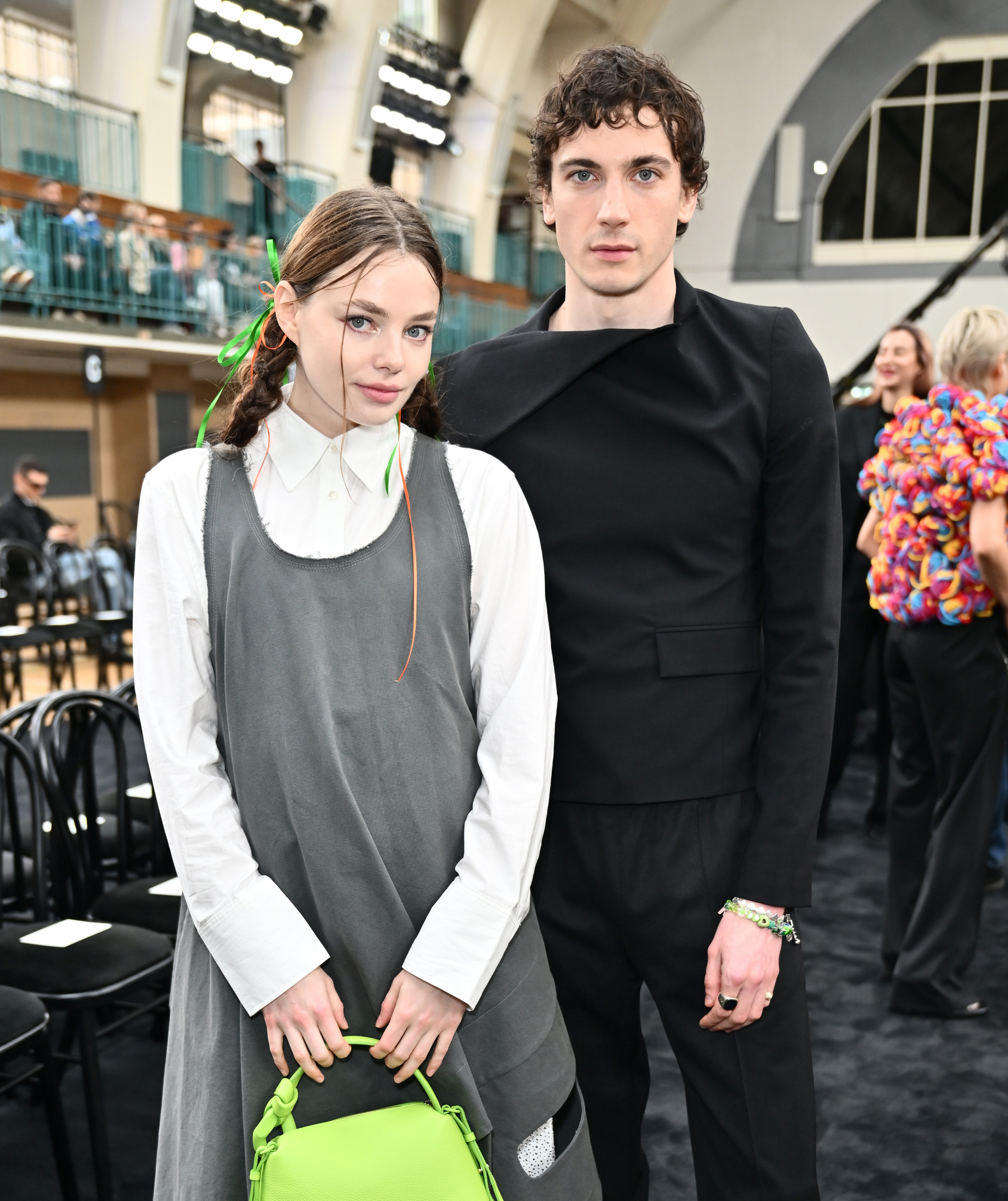 Two individuals at an event, one in a grey dress with a green accent and a neon bag, the other in a black outfit