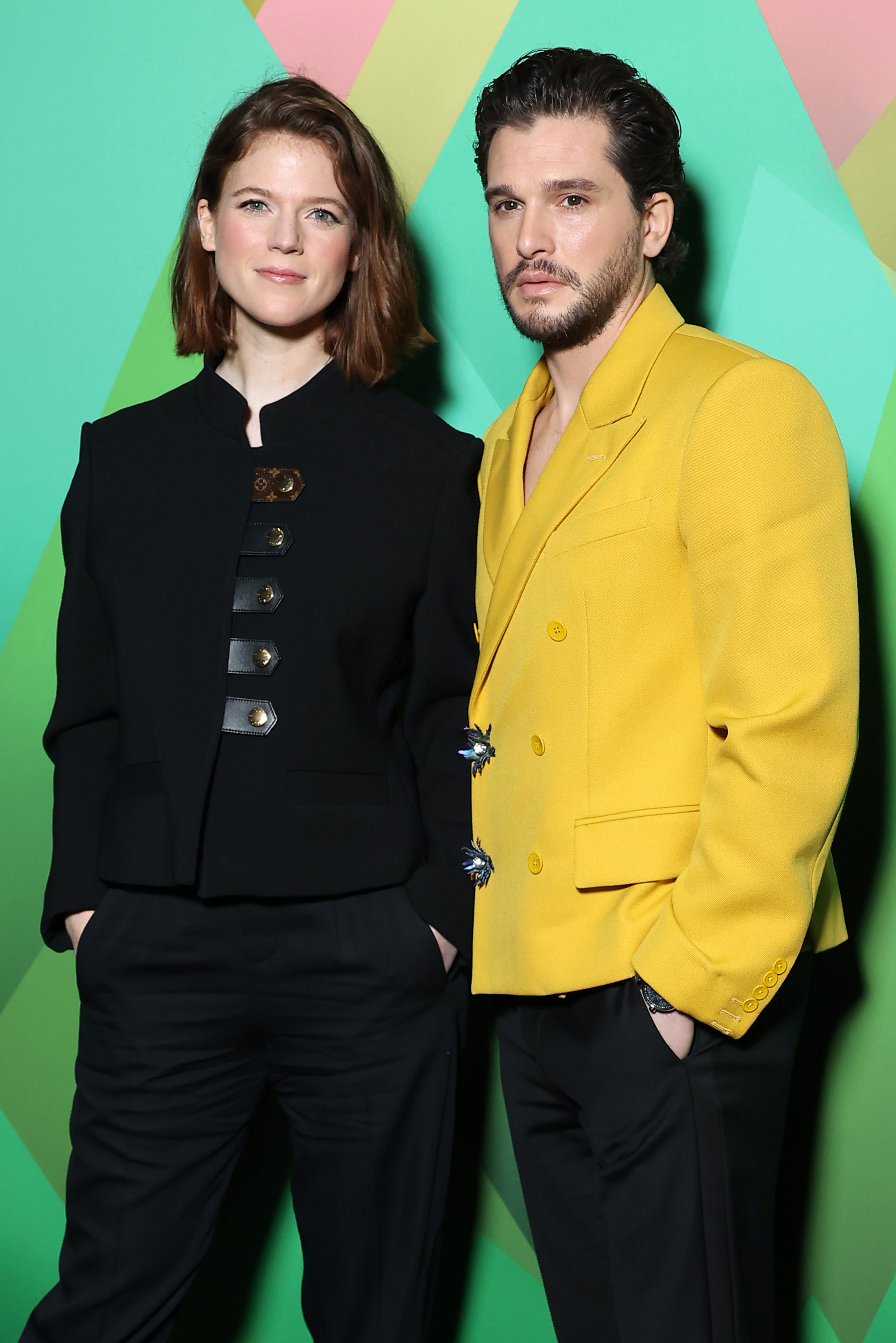 Two individuals posing, one in a black outfit with detailed buttons, the other in a bright yellow suit jacket