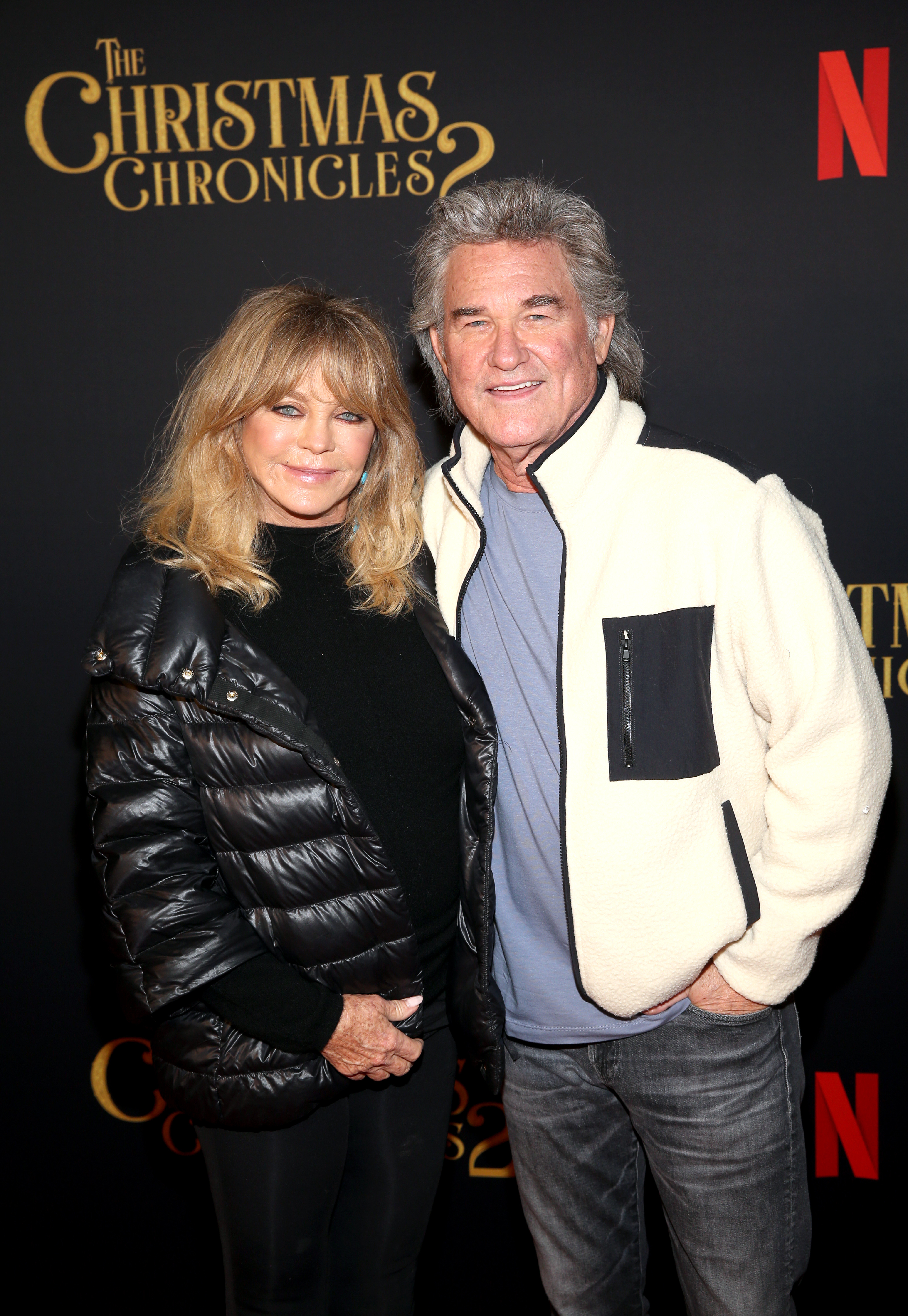 Two celebrities pose together; one in a black puffer jacket, the other in a cream jacket with a dark pocket detail