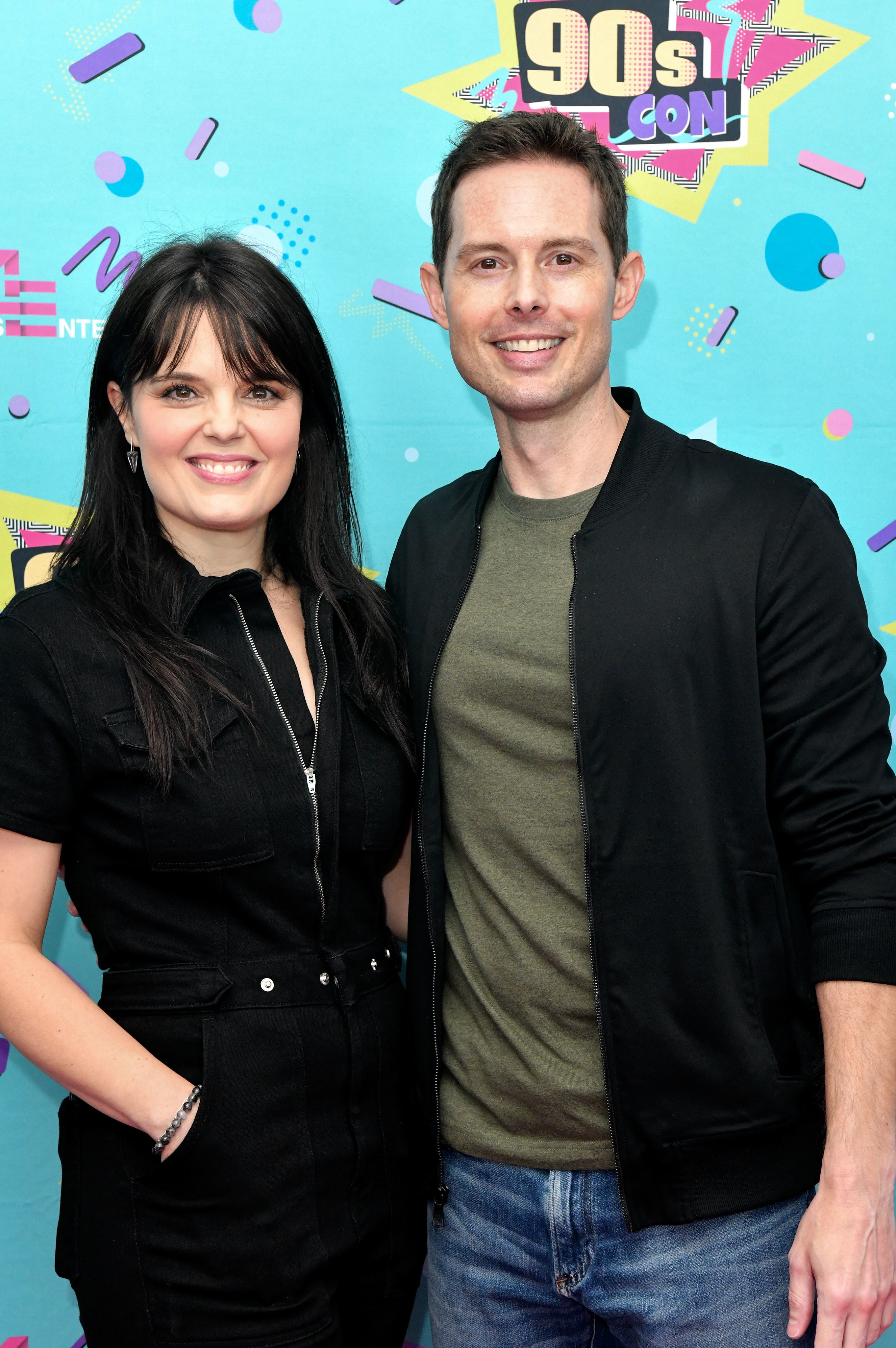 Two individuals posing together at a &#x27;90s Con event, woman in black outfit, man in black jacket and green shirt