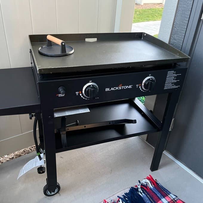 Flat top outdoor griddle with a spatula on it, under a patio, next to a folded blanket. Brand is &quot;Blackstone.&quot;