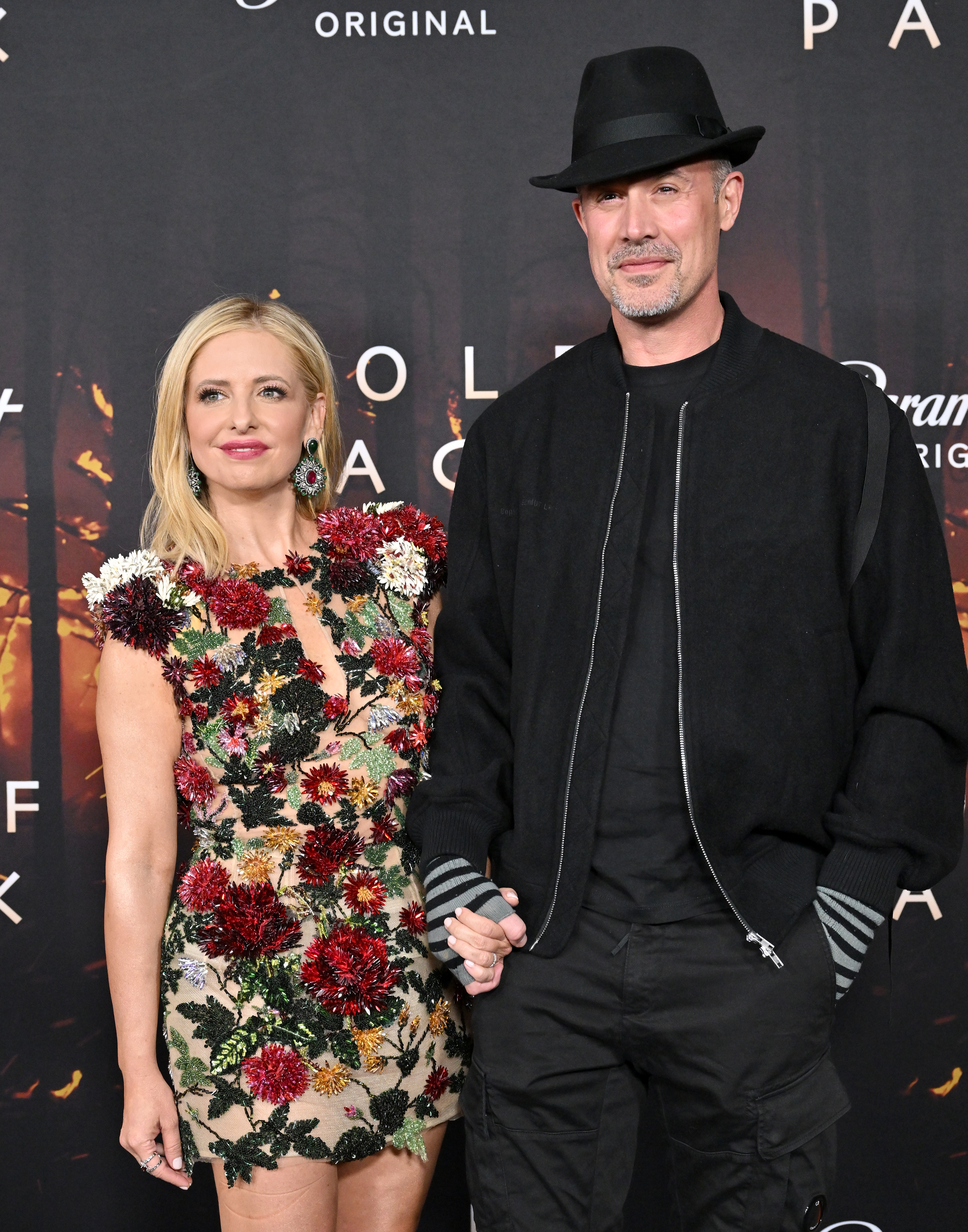 Two individuals posing together; one in a floral embellished dress and the other in a black outfit with a hat