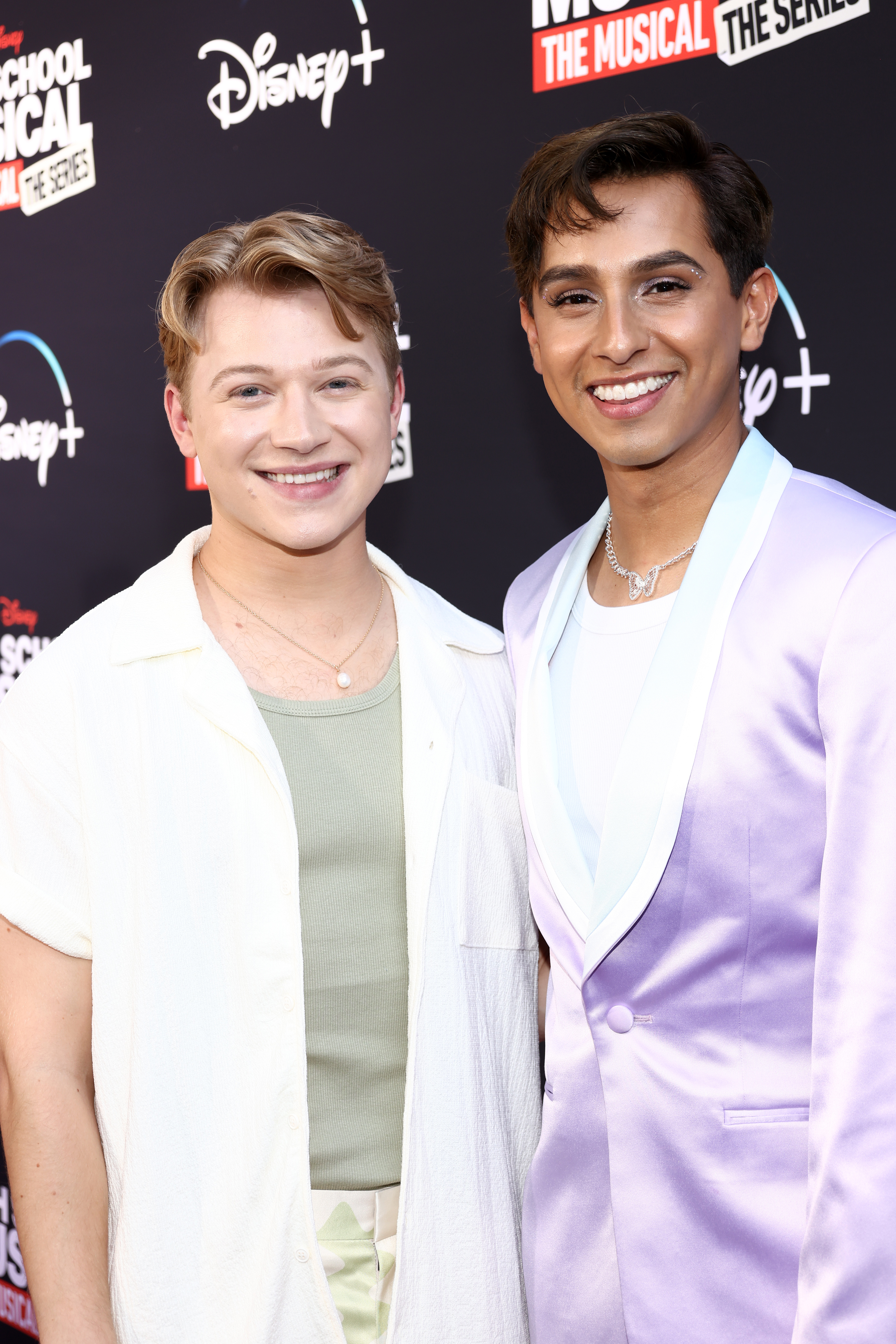 Two actors from &quot;High School Musical: The Musical&quot; smiling at a promotional event. One wears a white outfit, the other a lavender suit
