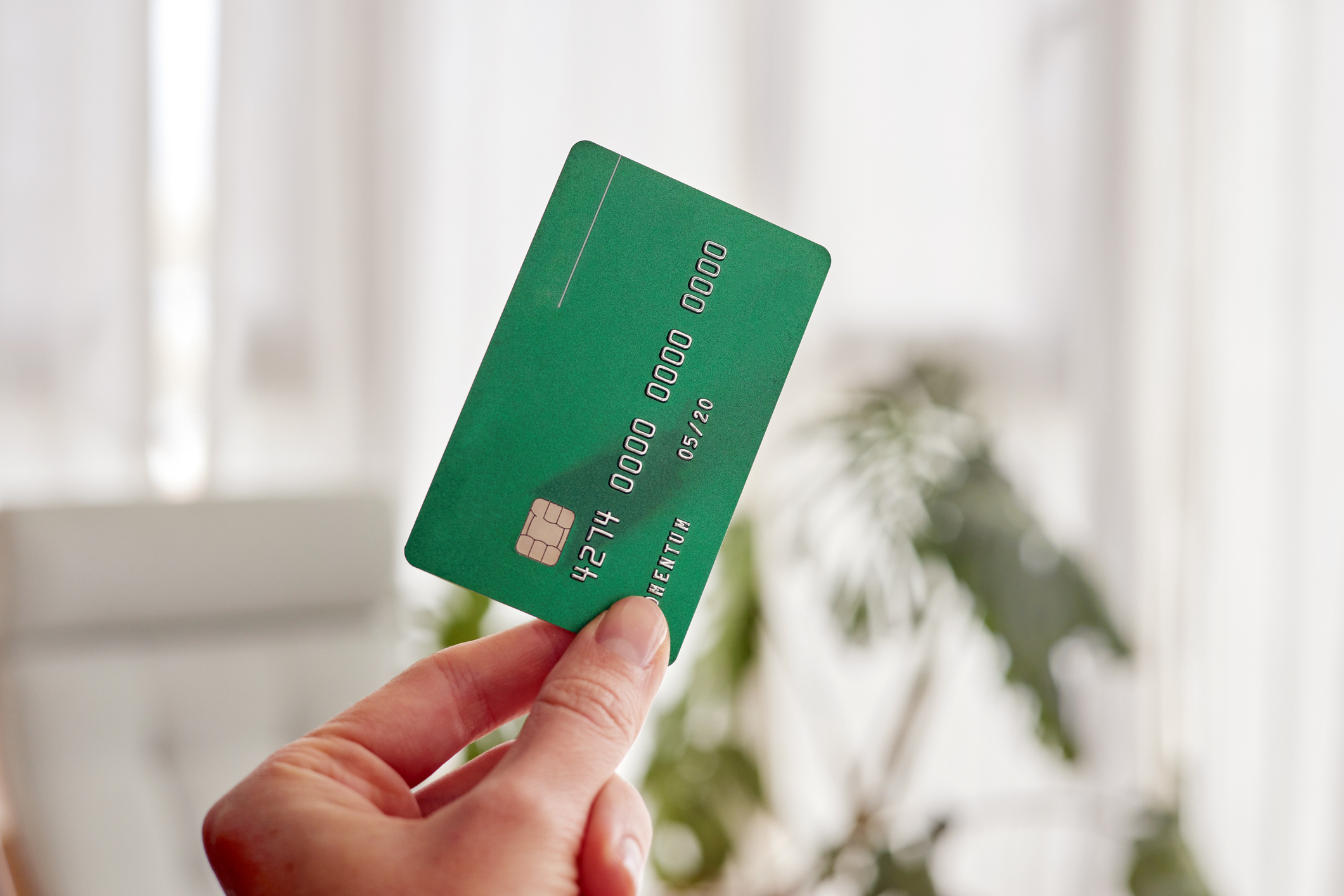Hand holding a green bank card indoors, partial view of a window in the background
