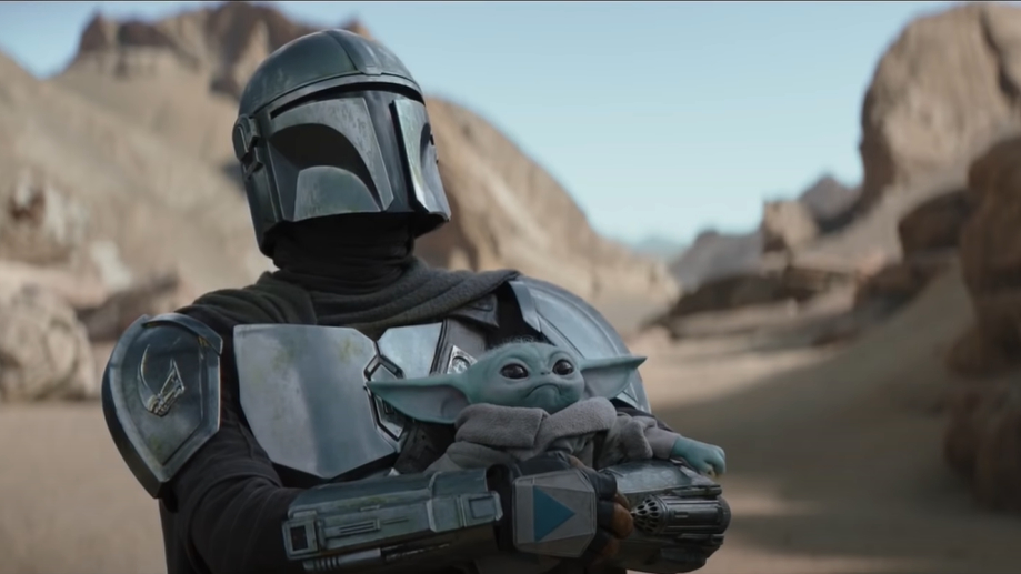The Mandalorian in armor holds Baby Yoda (Grogu) in a desert-like terrain