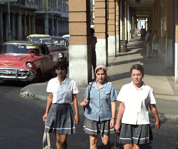 Three women in skirts and shirts walking on a sidewalk with vintage cars in background