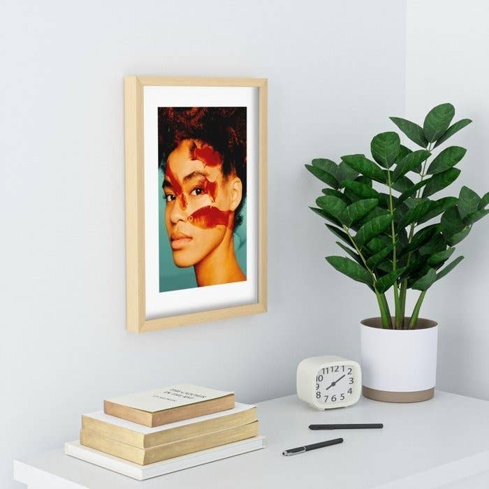 Framed portrait of an unidentifiable woman hanging on a wall next to a potted plant and books on a shelf