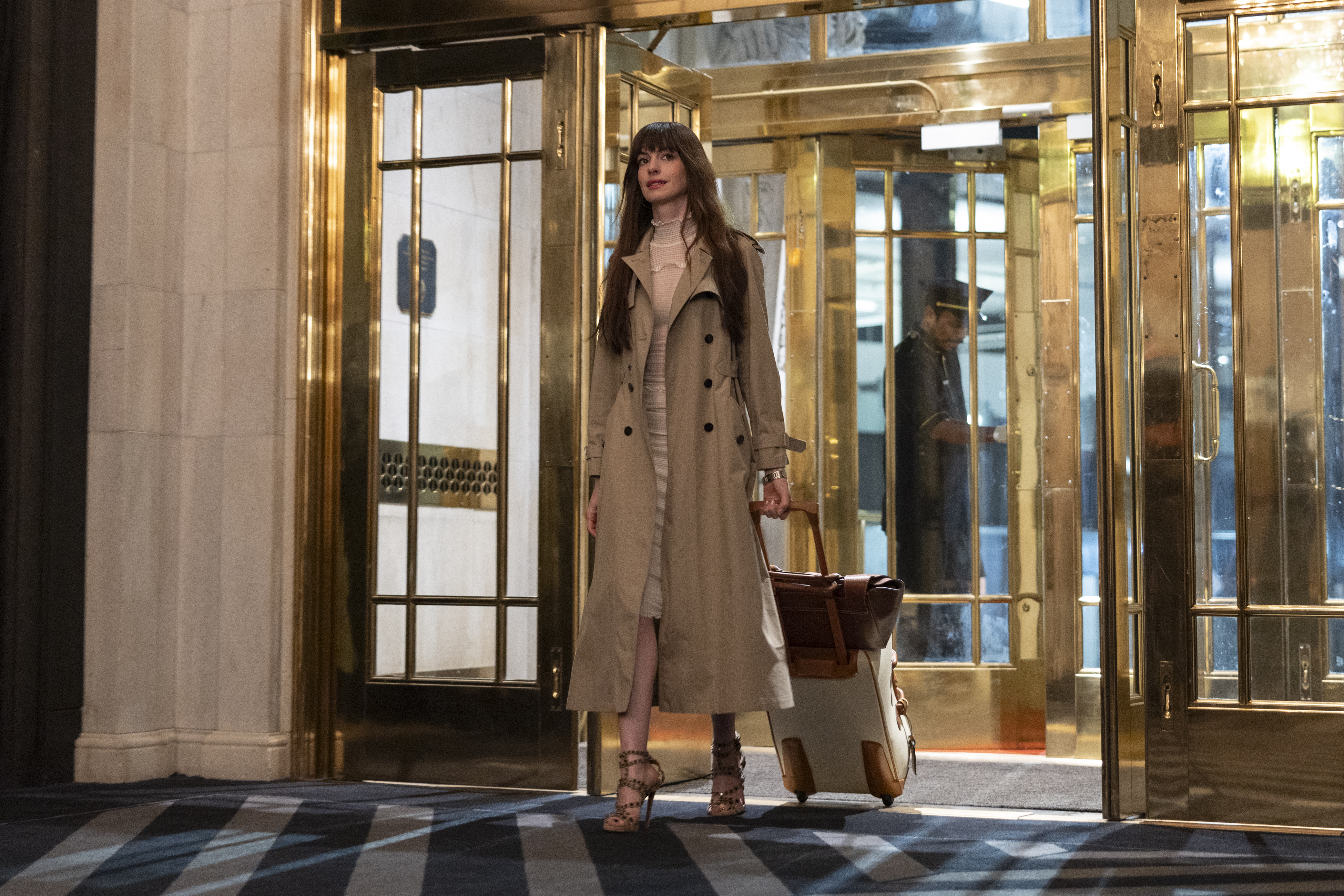 Woman in a trench coat carrying luggage exits a building, in what appears to be a scene from a film or TV show