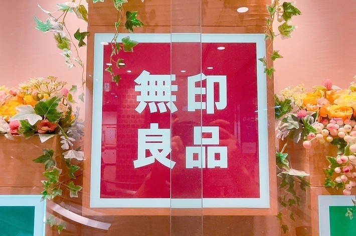 画像は「無印良品」と書かれた赤い看板です。花飾りに囲まれています。