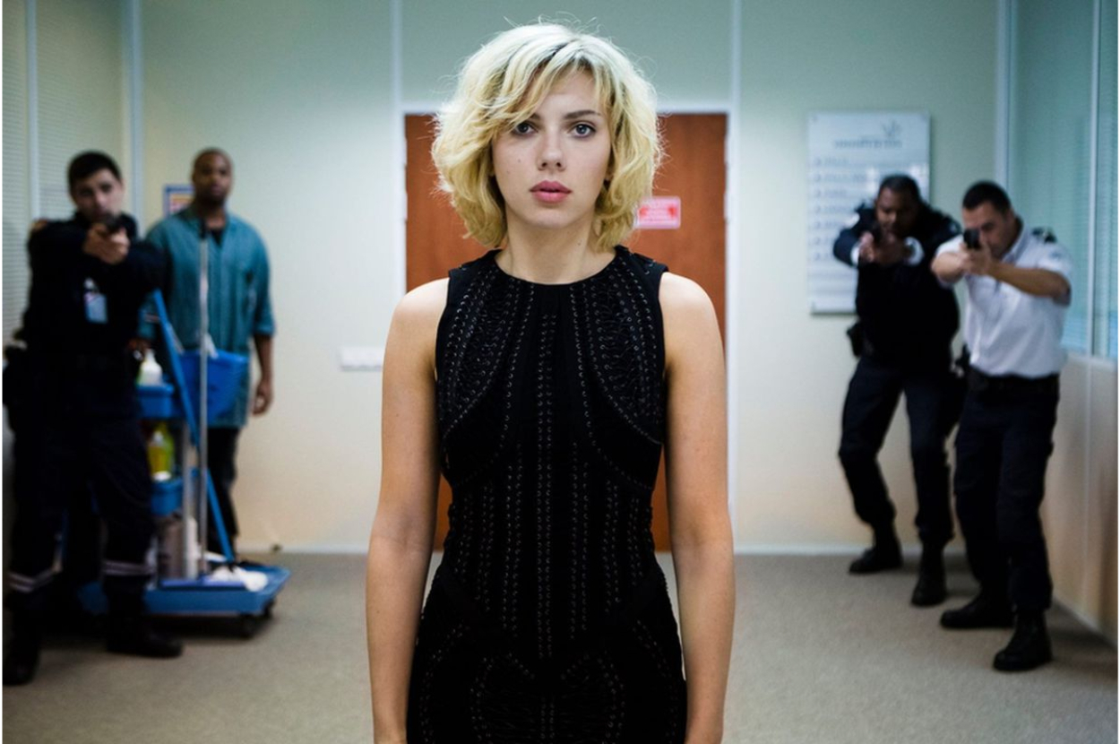 Scarlett Johansson in character, wearing a sleeveless black dress, with actors portraying security personnel in the background