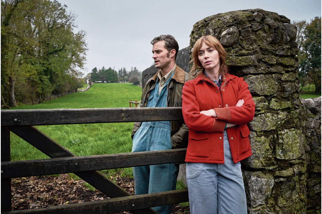 Two actors posing against a fence in a rural setting, dressed in casual jackets and jeans