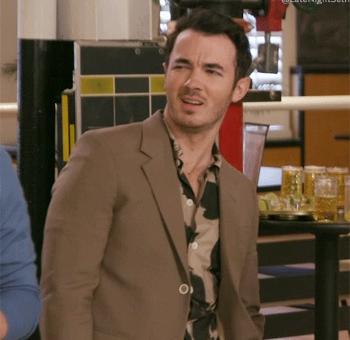 Kevin Jonas at Late Night with Seth Meyers set, reacting with a confused expression while in a conversation; stylishly dressed in a brown blazer over a patterned shirt