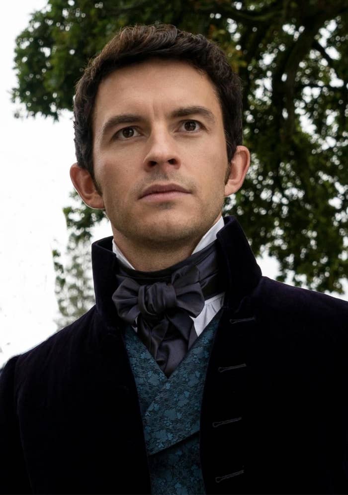 Jonathan Bailey in period costume with a high-collared coat and cravat, in a scene from a TV show or movie set against an outdoor backdrop with greenery