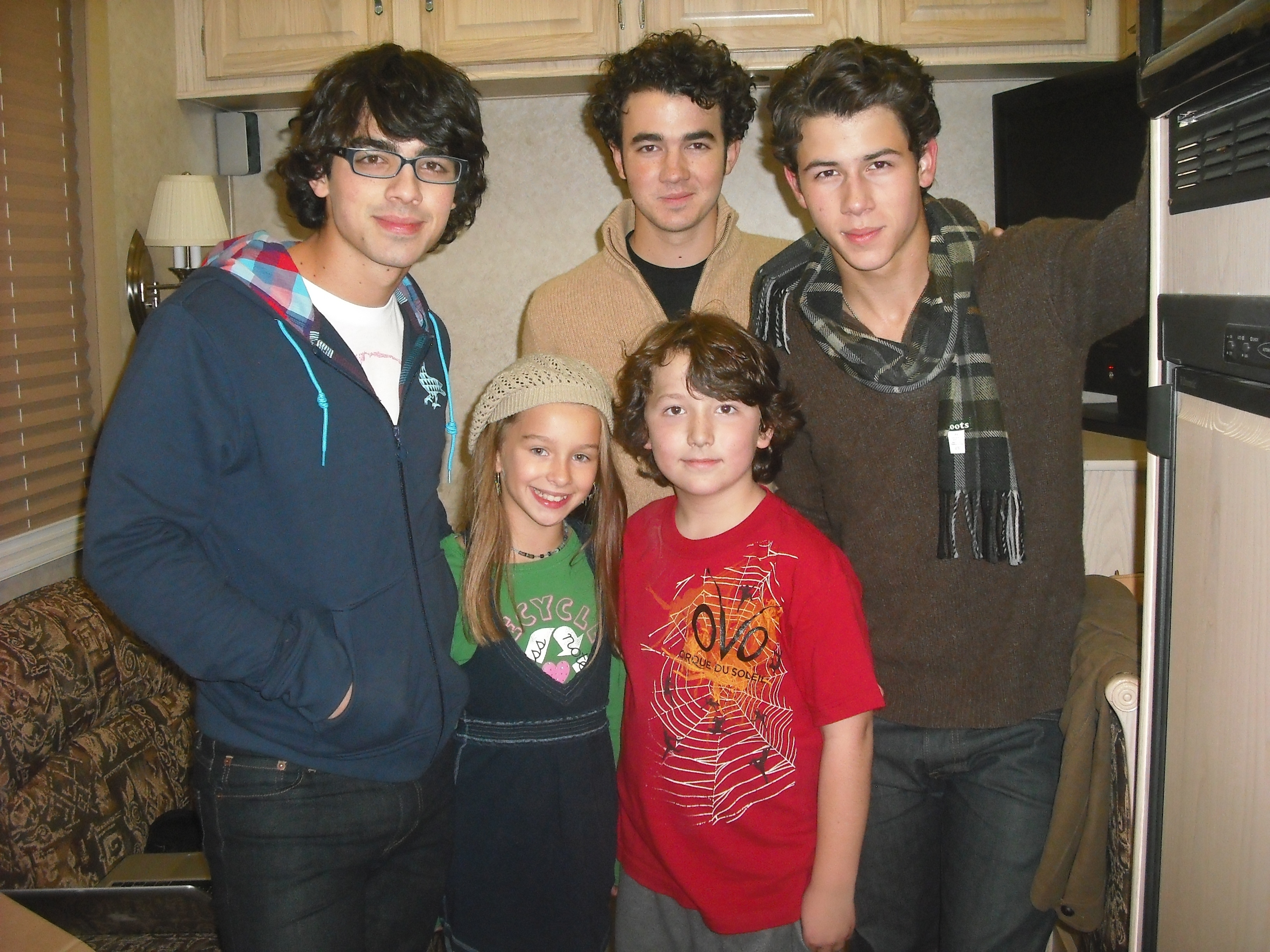 Joe Jonas, Kevin Jonas, Nick Jonas, and two children smile for a photo indoors. Joe wears glasses and a hoodie; Nick has a scarf