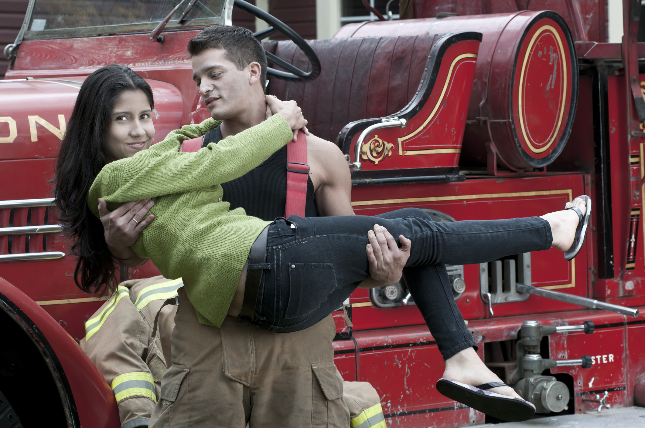 Man in a fireman uniform playfully carries a smiling woman wearing a green sweater and jeans in front of a vintage fire truck