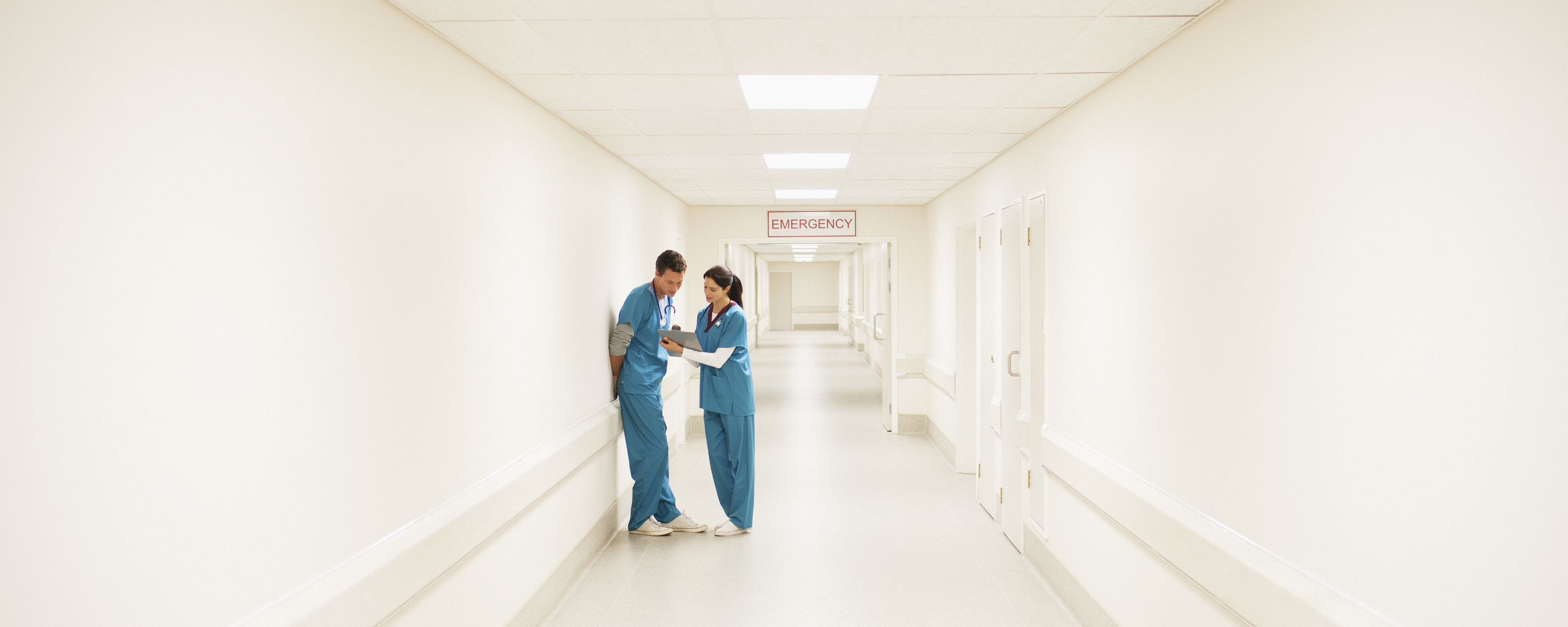 Two medical professionals in blue scrubs stand in a hospital hallway, engaged in conversation near an &quot;Emergency&quot; sign at the hallway&#x27;s end