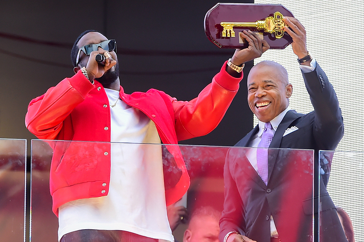 Sean "Diddy" Combs holds a microphone and wears a casual outfit with a red jacket, standing beside a man in a suit who holds a ceremonial key on stage