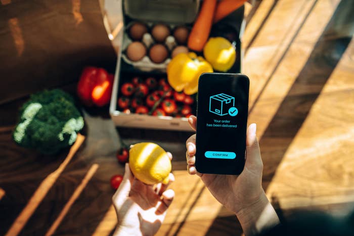 Person holding a lemon in one hand and a smartphone showing "Your order has been delivered" in the other, with various groceries on a table from a subscription box