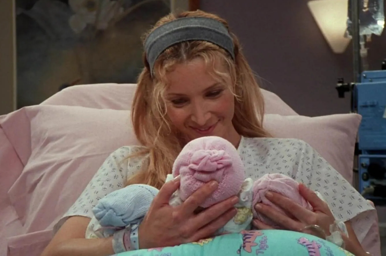 Lisa Kudrow, in a hospital gown, holding three newborn babies wrapped in blankets, smiling while looking at them