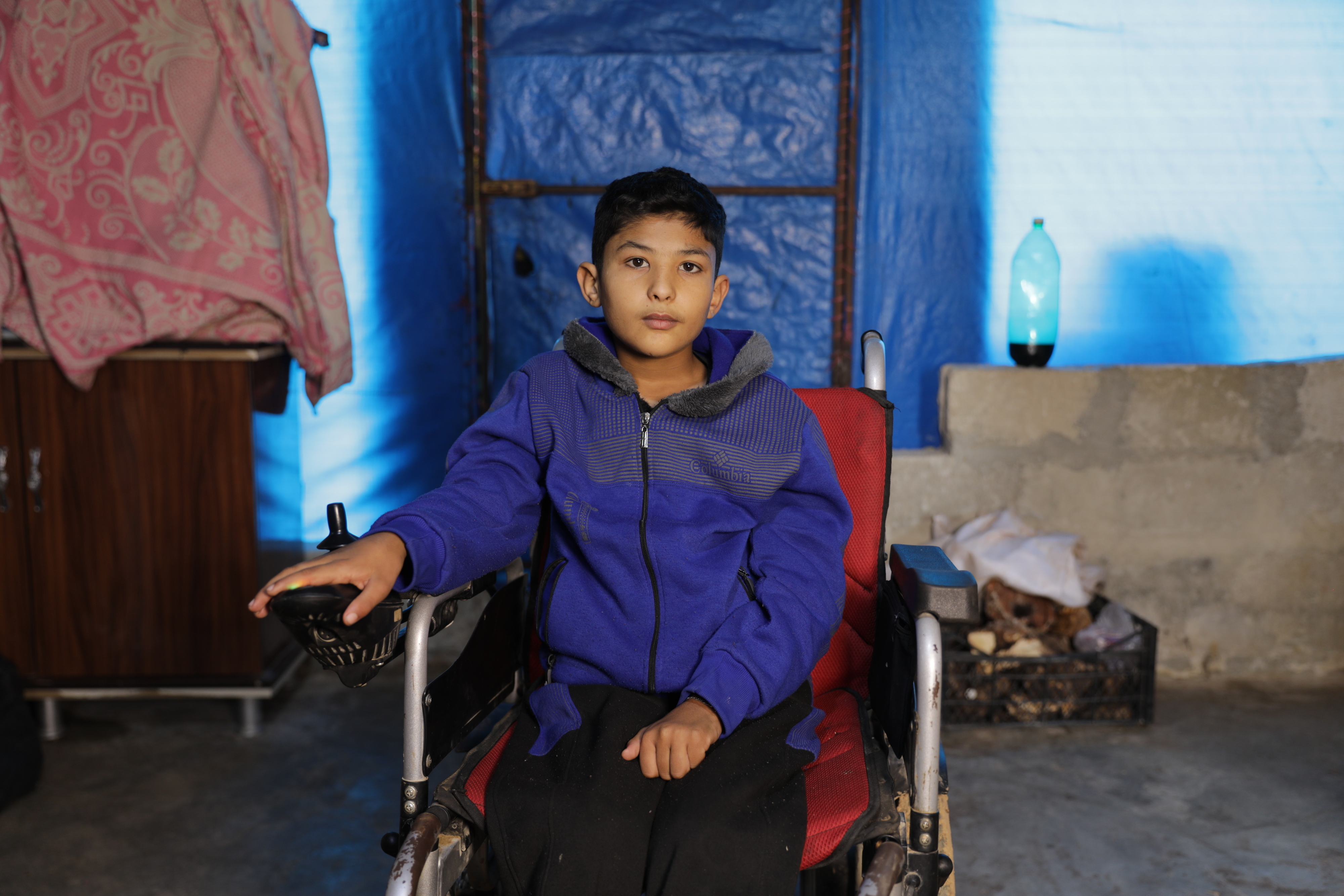 A young boy sits in a wheelchair in a modest, makeshift dwelling; he wears a blue jacket and black pants, with a thoughtful expression on his face