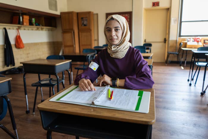 A woman, wearing a modest outfit and hijab, sits alone at a desk in a classroom with papers and a pen on her desk