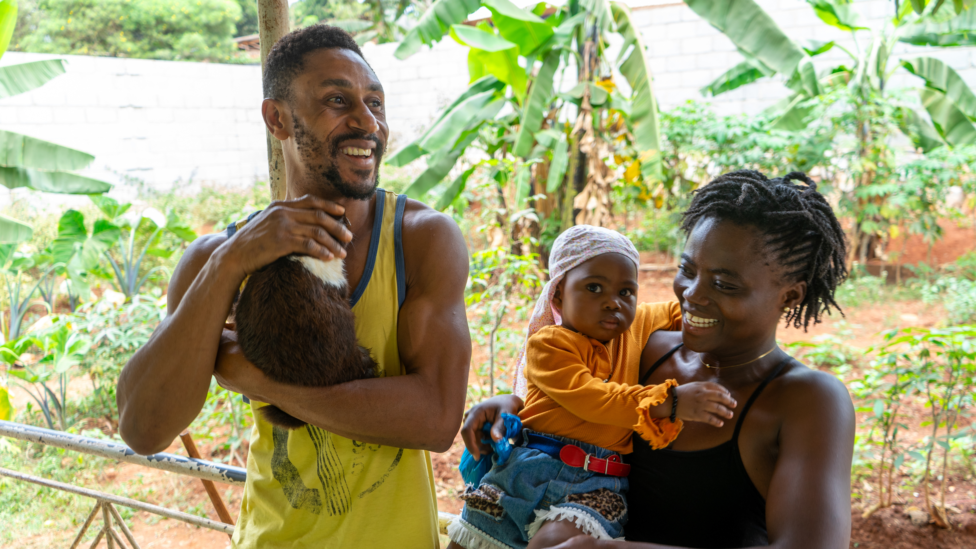 A man holds a small animal while smiling at a woman holding a baby; they stand outside near green foliage and appear happy and relaxed
