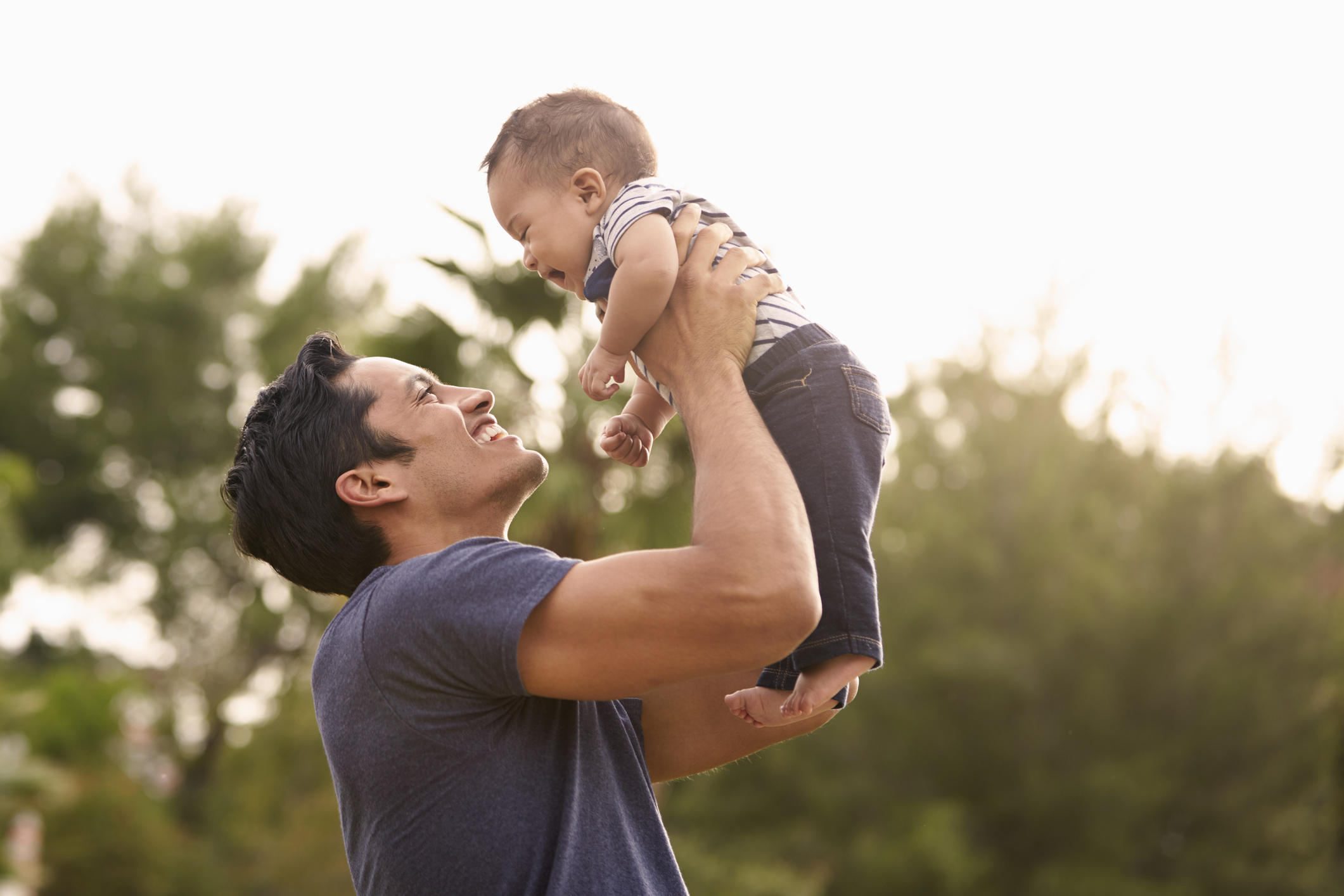 A smiling man in a plain t-shirt holds up a happy baby with both hands outdoors