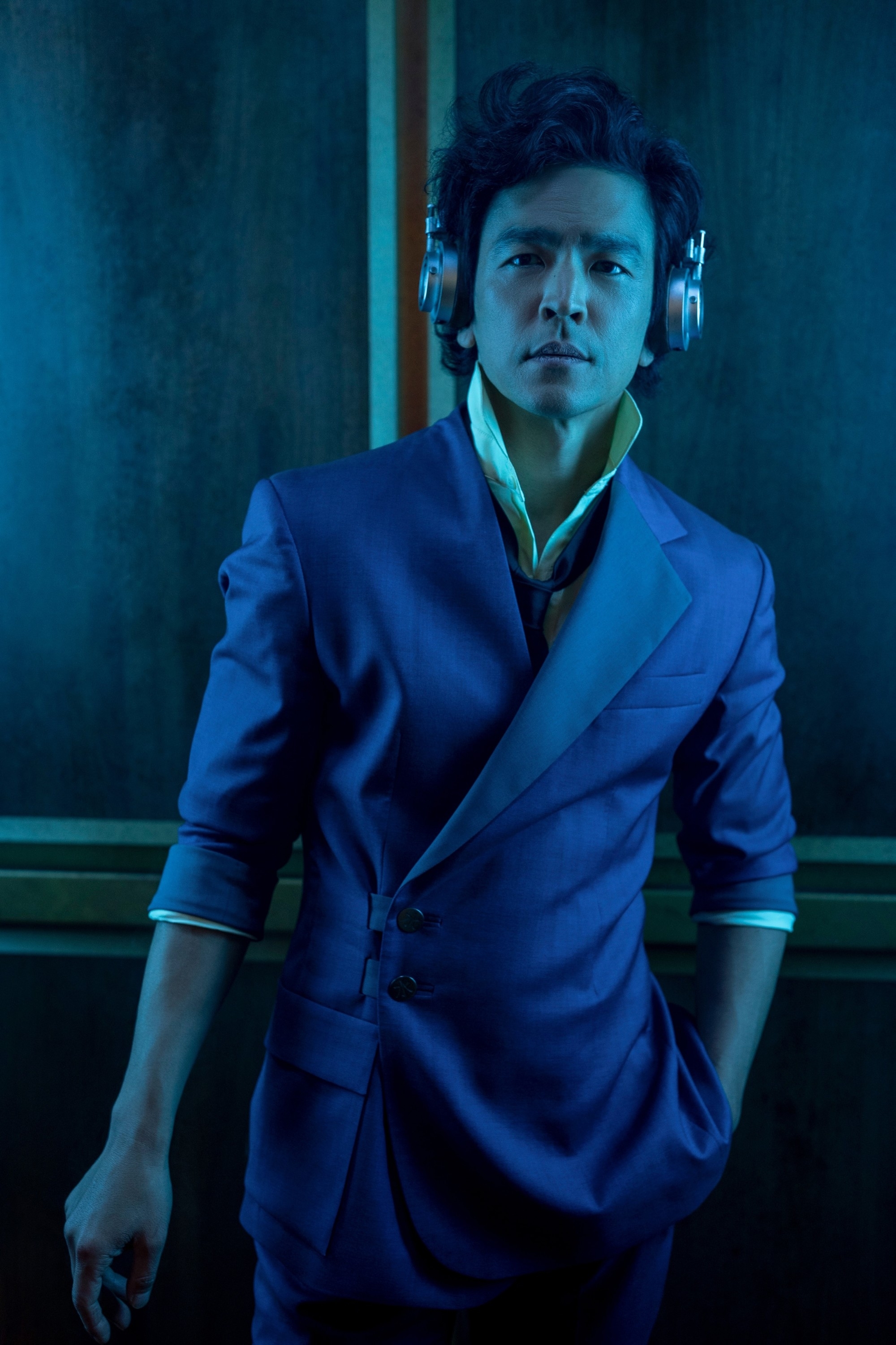 John Cho, wearing a modern, asymmetrical suit and headphones, poses against a dark, indoor backdrop