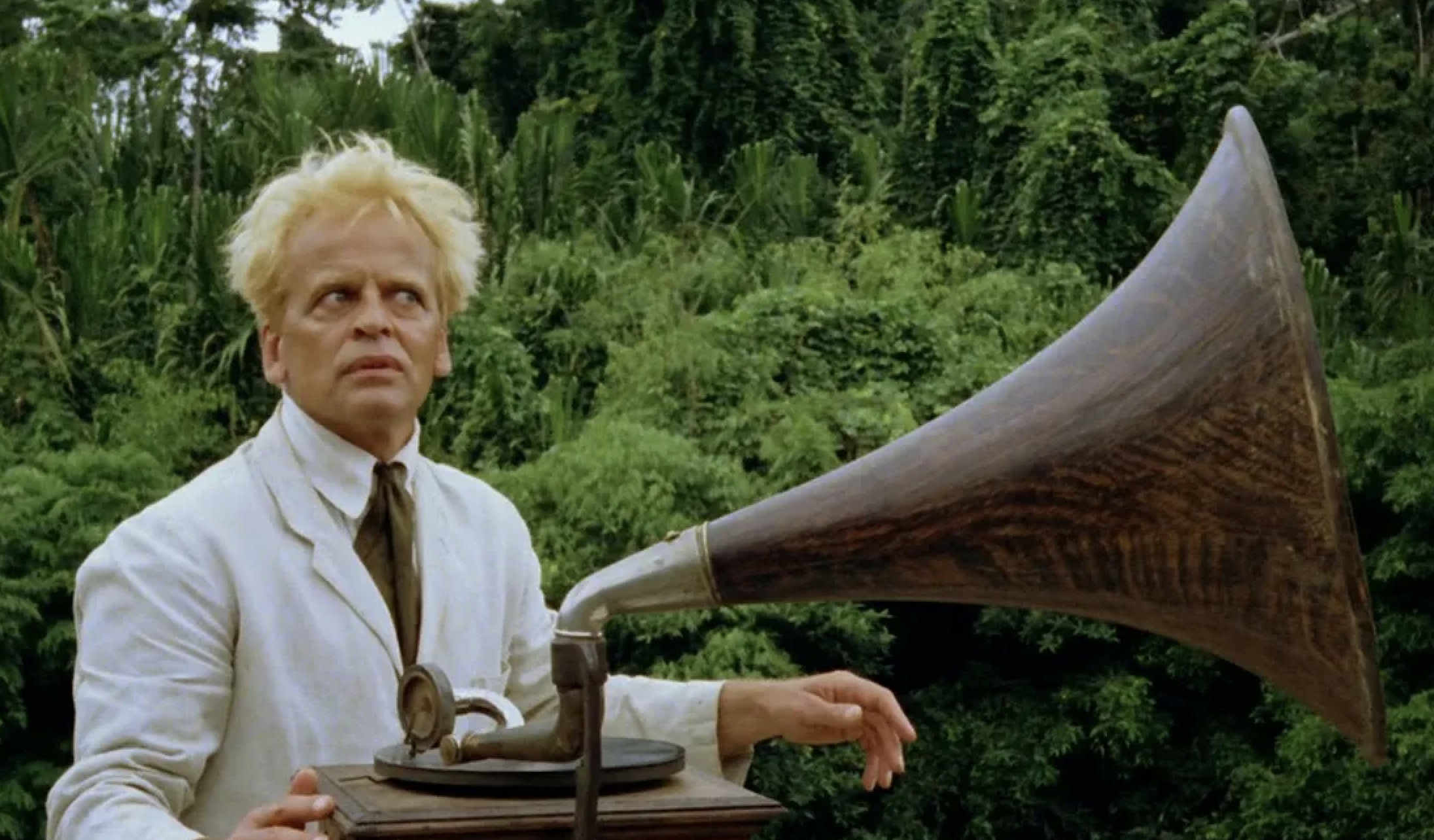 A man with wild blond hair, dressed in a white suit and tie, stands in a lush forest with an antique phonograph