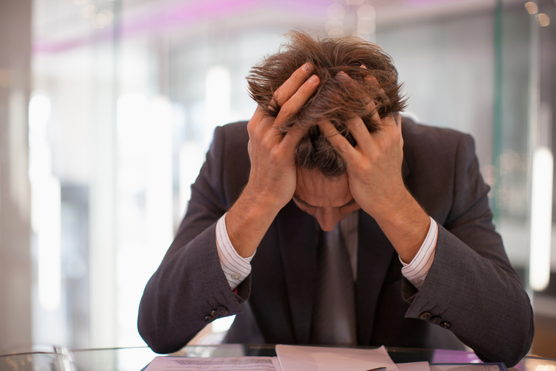 A person in a suit is sitting at a desk with their head in their hands, appearing stressed while looking down at documents