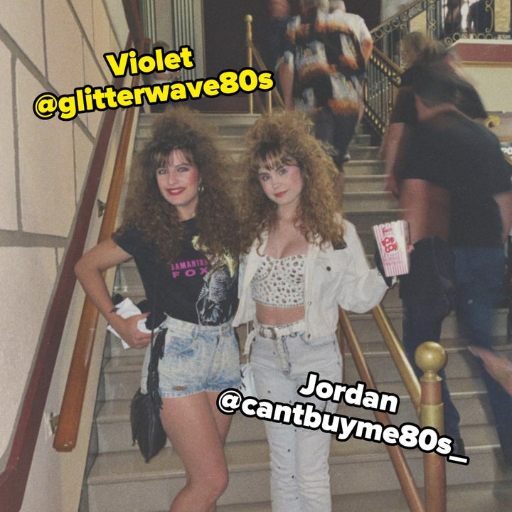 Two women with curly hair, dressed in casual 1980s fashion, stand on stairs; one in a black graphic T-shirt and denim shorts, the other in white jeans and a crop top